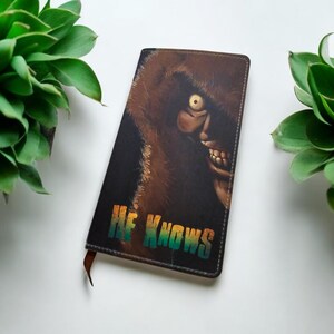 May include: A brown leather notebook with a graphic of a creature with a single eye and a toothy grin. The text "HE KNOWS" is printed in rainbow colors on the front cover.