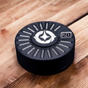 May include: A black and white circular device with a white starburst design and the number 20 on a small white square. The device is on a wooden surface.