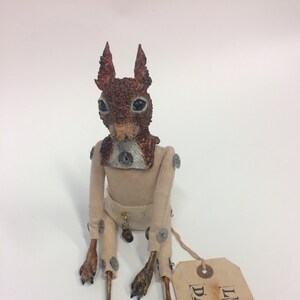May include: A handmade doll with a squirrel head, a beige fabric body, and articulated limbs. The doll is wearing a white fabric onesie with metal fasteners. The doll is sitting on a white surface with a brown paper tag that reads "LITTLE DAEMON".