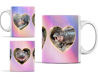 May include: A white ceramic mug with a pink and purple ombre background. The mug features three images of a baby hippopotamus in heart-shaped frames with a gold outline. The hippopotamus is in different poses, including yawning and smiling.