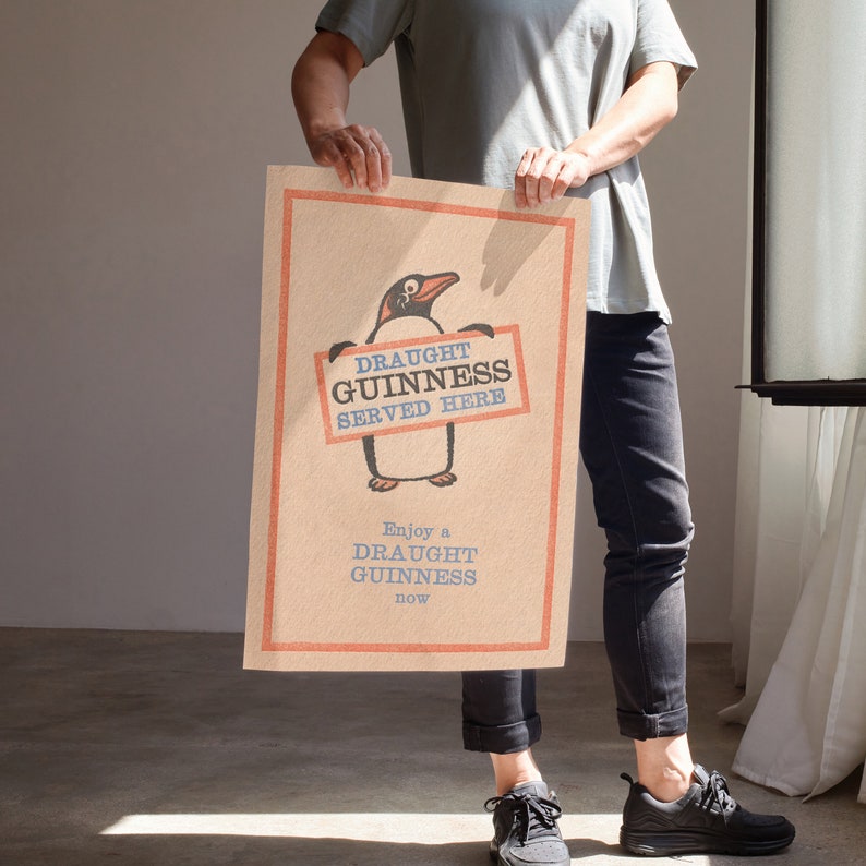 Puede incluir: Un p&oacute;ster de estilo vintage con un ping&uuml;ino de dibujos animados sosteniendo un cartel que dice "Draught Guinness Served Here". El p&oacute;ster tiene un fondo marr&oacute;n con acentos rojos y azules. El texto "Enjoy a Draught Guinness now" est&aacute; impreso debajo del ping&uuml;ino.