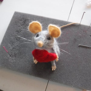 Puede incluir: Un rat&oacute;n de fieltro gris que sostiene un coraz&oacute;n de fieltro rojo. El rat&oacute;n tiene orejas beige y ojos negros. El rat&oacute;n est&aacute; sentado sobre una superficie de espuma gris.