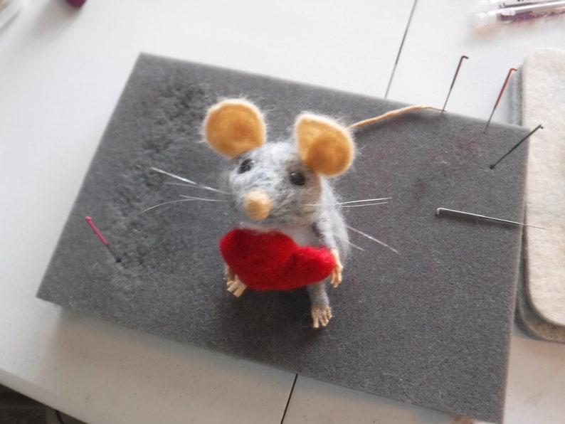 Puede incluir: Un rat&oacute;n de fieltro gris que sostiene un coraz&oacute;n de fieltro rojo. El rat&oacute;n tiene orejas beige y ojos negros. El rat&oacute;n est&aacute; sentado sobre una superficie de espuma gris.