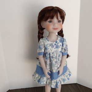May include: A doll with auburn hair styled in two braids, featuring blue eyes and rosy cheeks. The doll wears a short-sleeved dress with a white and blue floral pattern, accented by a solid blue horizontal band across the skirt and blue sleeve cuffs.