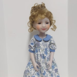 May include: A doll with curly blonde hair and a fair complexion wearing a dress featuring a blue and white floral pattern. The dress has short puff sleeves with blue cuffs and a rounded blue collar. The doll has a gentle expression with rosy cheeks.