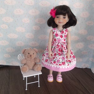 May include: A doll wearing a pink and white floral dress with a pink bow in her hair. She is standing next to a white chair with a brown teddy bear sitting on it.