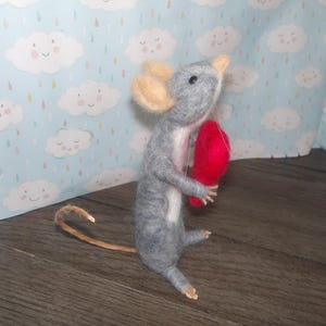 Puede incluir: Un rat&oacute;n de fieltro gris que sostiene un coraz&oacute;n de fieltro rojo. El rat&oacute;n tiene una cola larga y est&aacute; de pie sobre una superficie de madera.