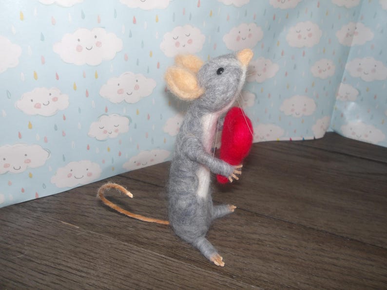 Puede incluir: Un rat&oacute;n de fieltro gris que sostiene un coraz&oacute;n de fieltro rojo. El rat&oacute;n tiene una cola larga y est&aacute; de pie sobre una superficie de madera.