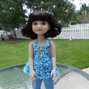 May include: A doll with dark hair wearing a blue floral print top with yellow buttons and blue pants. The doll is standing on a glass table in front of a white picket fence and green grass.