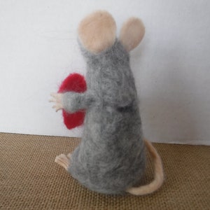 Puede incluir: Un rat&oacute;n de fieltro gris que sostiene un coraz&oacute;n de fieltro rojo. El rat&oacute;n tiene una cola larga y orejas rosas.
