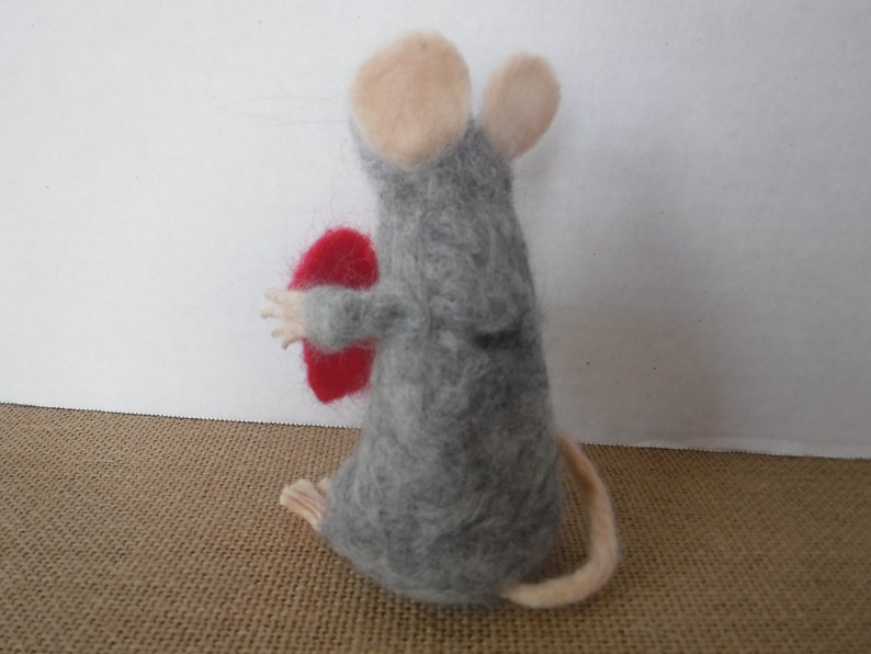 Puede incluir: Un rat&oacute;n de fieltro gris que sostiene un coraz&oacute;n de fieltro rojo. El rat&oacute;n tiene una cola larga y orejas rosas.