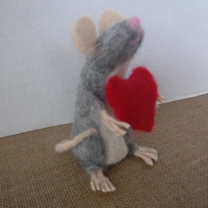 Puede incluir: Un rat&oacute;n de fieltro gris que sostiene un coraz&oacute;n de fieltro rojo. El rat&oacute;n tiene la nariz rosa y bigotes blancos.