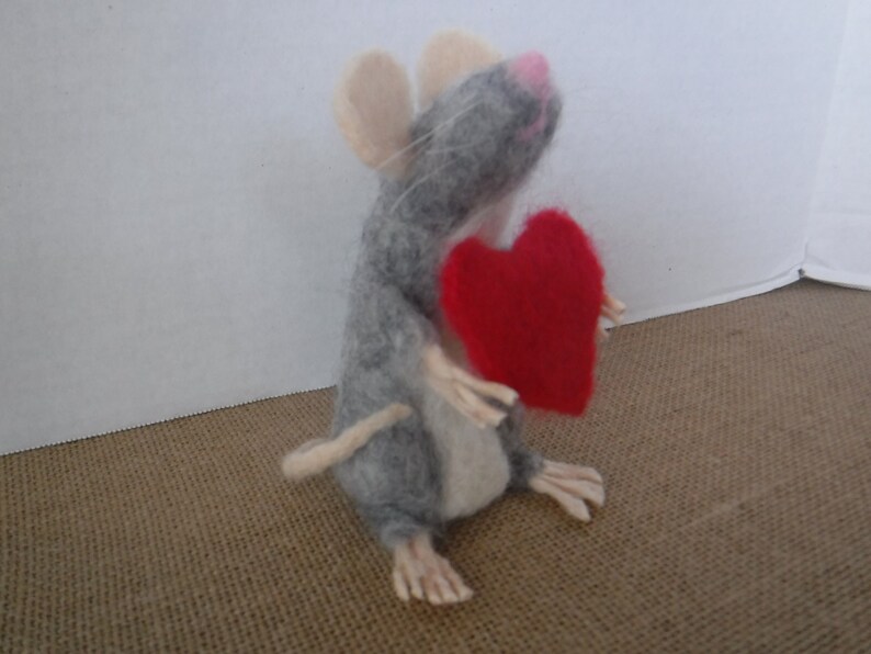 Puede incluir: Un rat&oacute;n de fieltro gris que sostiene un coraz&oacute;n de fieltro rojo. El rat&oacute;n tiene la nariz rosa y bigotes blancos.