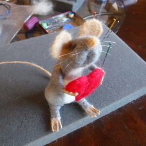 Puede incluir: Un rat&oacute;n de fieltro de aguja gris y blanco sosteniendo un coraz&oacute;n de fieltro rojo. El rat&oacute;n est&aacute; sentado sobre una superficie de espuma gris.