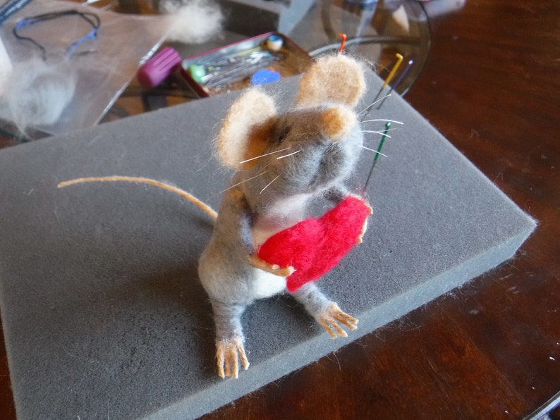 Puede incluir: Un rat&oacute;n de fieltro de aguja gris y blanco sosteniendo un coraz&oacute;n de fieltro rojo. El rat&oacute;n est&aacute; sentado sobre una superficie de espuma gris.