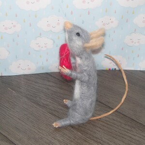 Puede incluir: Un rat&oacute;n de fieltro gris que sostiene un coraz&oacute;n de fieltro rojo. El rat&oacute;n tiene una cola larga y est&aacute; de pie sobre una superficie de madera.
