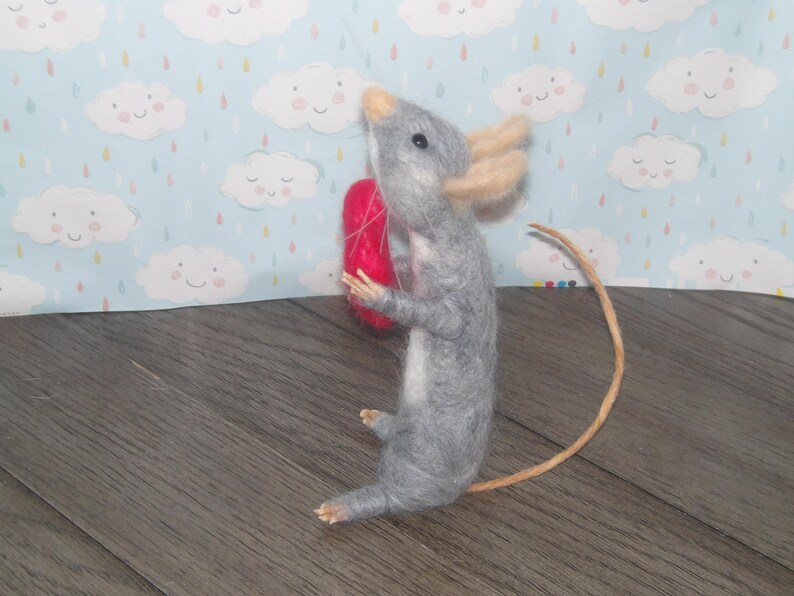 Puede incluir: Un rat&oacute;n de fieltro gris que sostiene un coraz&oacute;n de fieltro rojo. El rat&oacute;n tiene una cola larga y est&aacute; de pie sobre una superficie de madera.