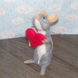Puede incluir: Un rat&oacute;n de fieltro gris y blanco sosteniendo un coraz&oacute;n de fieltro rojo. El rat&oacute;n tiene una cola larga y est&aacute; de pie sobre una superficie de madera.