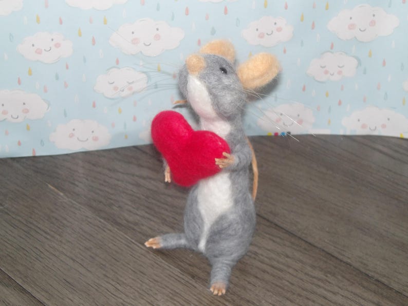 Puede incluir: Un rat&oacute;n de fieltro gris y blanco sosteniendo un coraz&oacute;n de fieltro rojo. El rat&oacute;n tiene una cola larga y est&aacute; de pie sobre una superficie de madera.
