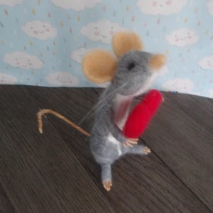 Puede incluir: Un rat&oacute;n de fieltro gris que sostiene un coraz&oacute;n de fieltro rojo. El rat&oacute;n tiene una cola larga y delgada y un par de orejas grandes y redondas.