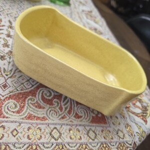 May include: A yellow ceramic dish with an elongated, rounded rectangular shape and a smooth, matte finish. The vessel features high, curved walls and a deep interior basin, presenting a solid, monochromatic color throughout its entire structure.