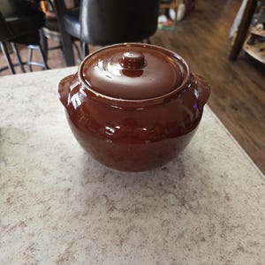 May include: A brown ceramic casserole dish with a lid. The dish has two small handles on the sides and a knob on the lid. The dish is sitting on a light-colored countertop. The casserole dish is a kitchenware item.