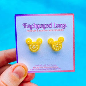 May include: A pair of yellow lemon-shaped earrings on a white card with the text "Enchanted Luna Fandom & Fantasy Accessories". The earrings are in the shape of a mouse head with a lemon slice design. The card has a purple and pink border.