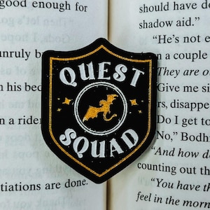 Pode incluir: Um patch preto e dourado em forma de escudo com as palavras "QUEST SQUAD" em branco e uma silhueta de dragão dourado. O patch está sobre as páginas de um livro aberto.