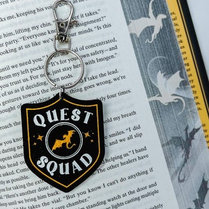 May include: Black and gold shield-shaped keychain with the text "QUEST SQUAD" in white, a gold dragon illustration, and star accents. The keychain has a silver clasp and ring, perfect for attaching to keys or a bag.