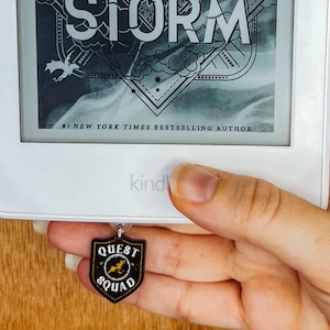 Könnte beinhalten: Ein weißer Kindle-E-Reader mit dem Buch "Onyx Storm" von Rebecca Yarros. Ein schwarzer und goldener "Quest Squad"-Anhänger hängt daran. Der Hintergrund ist eine holzfarbene Oberfläche.