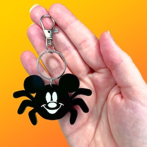 May include: Black Mickey Mouse spider keychain with a silver clasp and ring. The spider has a smiling face and eight legs. The keychain is held against a gradient orange and yellow background.