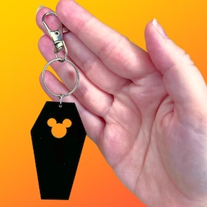 May include: Black coffin-shaped keychain with a silver clasp and ring. The keychain features a cut-out of a mouse head. The background is a gradient of orange and yellow.