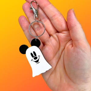 May include: A Halloween-themed keychain featuring a white ghost with a smiling face and black Mickey Mouse ears. The keychain has a silver clasp and a circular ring. The background is a gradient of orange and yellow.