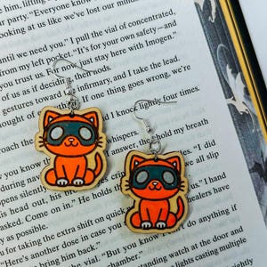 May include: A pair of orange cat-shaped earrings with black and green masks. The earrings are made of wood and have silver-colored hooks. The cat design is outlined in black and has a smiling face. These earrings are a fun accessory.
