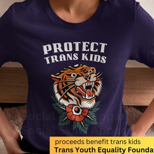 Può includere: Una maglietta blu navy con un'immagine di una tigre con un fiore. Il testo "PROTECT TRANS KIDS" è stampato sulla maglietta. Il testo "proceeds benefit trans kids Trans Youth Equality Foundation" è stampato su uno striscione giallo.