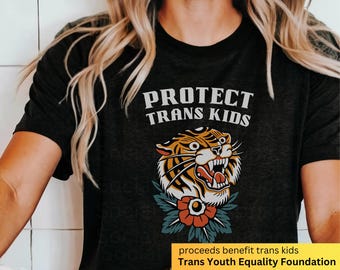Può includere: Maglietta nera con il testo "PROTECT TRANS KIDS" in lettere bianche. Un tigre di cartone animato con un fiore è stampato sotto il testo. Il testo "proceeds benefit trans kids Trans Youth Equality Foundation" è stampato in fondo alla maglietta.