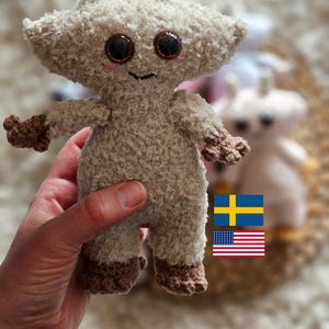 May include: A handmade, beige crochet doll with large, dark eyes and a smiling face. The doll has small arms and legs with brown accents. The doll is held in a hand, with a second doll visible in the background. The doll has two small antennae.