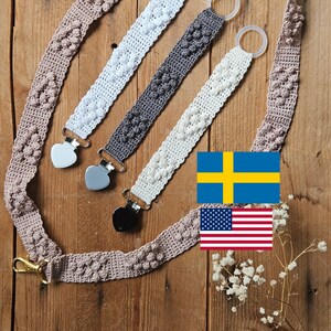 May include: A collection of handmade crochet pacifier clips in white, gray, and cream, with heart-shaped metal clips. A matching crochet strap with a gold-colored clasp is also present. The items are displayed on a wooden surface.