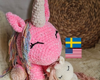 Puede incluir: Dos peluches de unicornio de crochet, uno rosa con una melena arcoíris y un cuerno blanco, y otro blanco con una melena rosa y un cuerno. El unicornio rosa es más grande y tiene los ojos cerrados. El unicornio blanco más pequeño está acostado con los ojos cerrados. Los peluches están sobre una tela de crochet blanca.