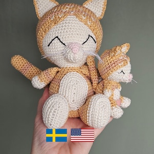 Puede incluir: Muñecas de gato de ganchillo en tonos beige, crema y rosa. El gato más grande tiene una cara sonriente con ojos negros y bigotes, y una versión más pequeña está sentada a su lado. Las muñecas están hechas a mano y tienen un aspecto suave y texturizado.