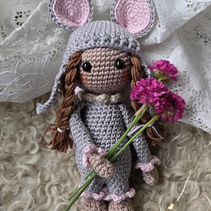 May include: A handcrafted crochet doll featuring a grey hooded outfit with pink-lined mouse ears, brown yarn hair styled in two braids, and a tan complexion. The doll holds a small bouquet of three vibrant magenta flowers with green stems.