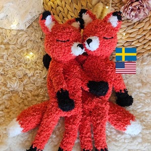 May include: Two plush red fox stuffed animals hugging each other. The foxes have black paws and ears, white accents, and closed eyes. The tails have white tips. The background includes a Swedish and American flag.