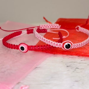 May include: Two red and pink string bracelets with an evil eye charm. The bracelets are on a white marble surface.