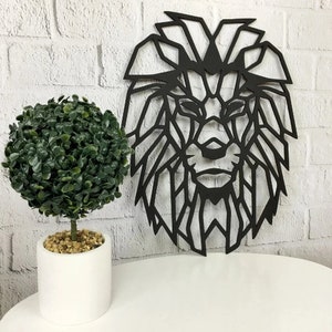 May include: A black geometric lion head wall art piece featuring sharp, angular lines and a cutout design. Beside it sits a small, round, green artificial topiary plant in a simple white cylindrical pot filled with small tan pebbles.