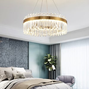 May include: A modern gold chandelier with clear crystal prisms hanging from a circular frame. The chandelier is in a bedroom setting with a bed, a chair, and a plant.
