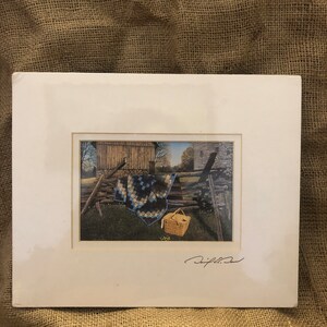 May include: A framed print featuring a rural scene with a wooden fence, a quilt, and a picnic basket. The quilt has a blue and black pattern. The print is in a white mat.