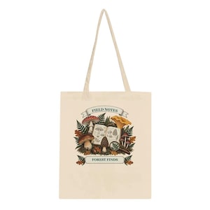 Op de afbeelding: Een beige canvas draagtas met een centrale afbeelding van diverse paddenstoelen, varens en een open veldnotitieboek. De tekst "FIELD NOTES" staat op een banner boven de illustratie en "FOREST FINDS" staat op een banner onder het ontwerp.