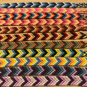 Può includere: Una collezione di braccialetti dell'amicizia colorati con motivi a chevron. I braccialetti sono realizzati con fili di diversi colori e sono legati insieme con nodi.