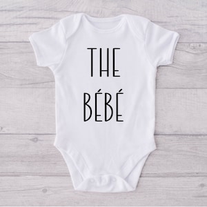 May include: White baby onesie with black text that reads "THE BÉBÉ".