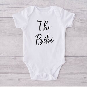 Può includere: Body da bambino bianco con maniche corte. Il testo "The Bébé" è scritto in corsivo nero sul davanti. Il body è realizzato in materiale morbido ed è progettato per neonati.